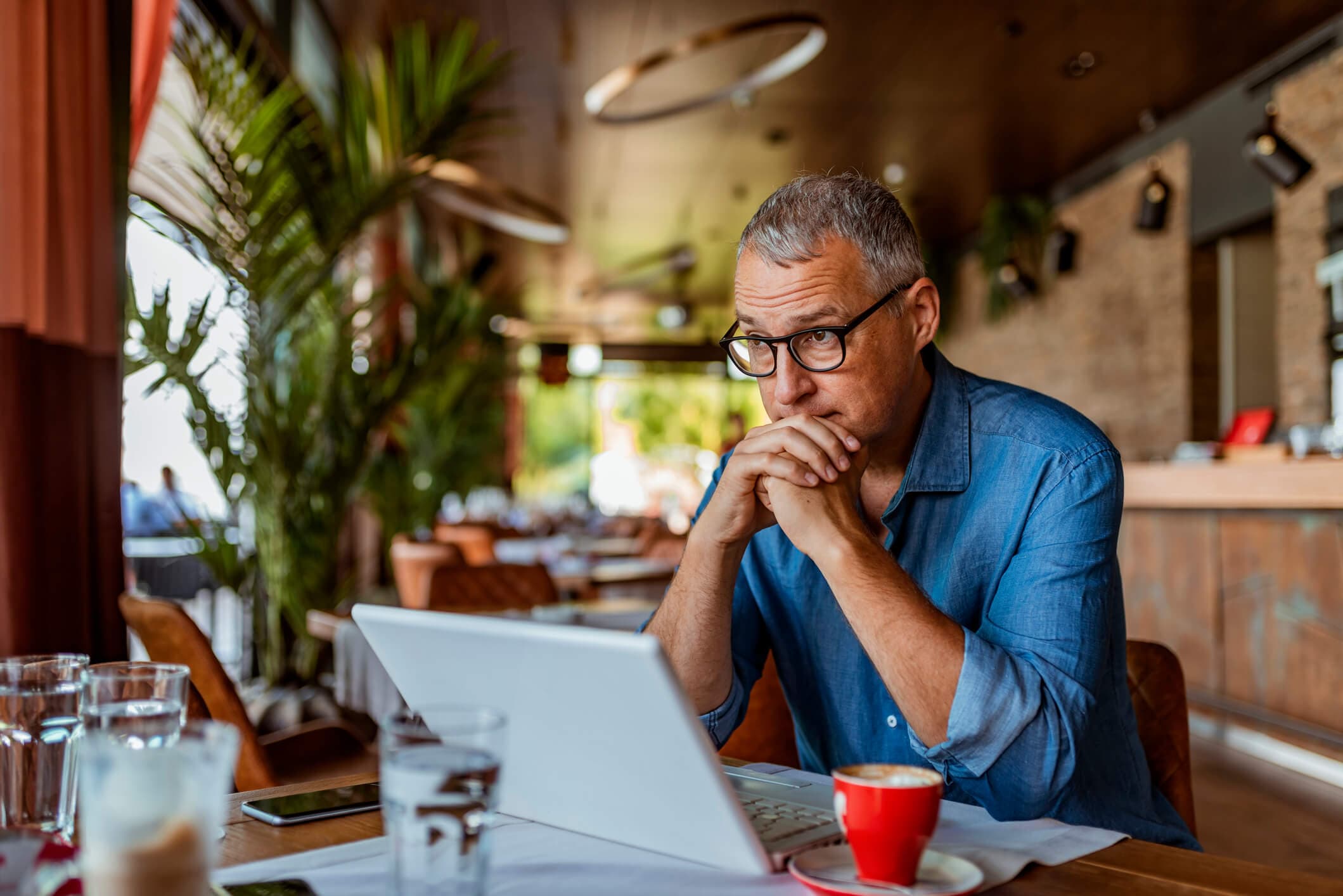 A middle-aged man sits at a table looking thoughtfully at his laptop, representing concerns about recession risk, retirement savings, investments, debt, and financial preparedness.