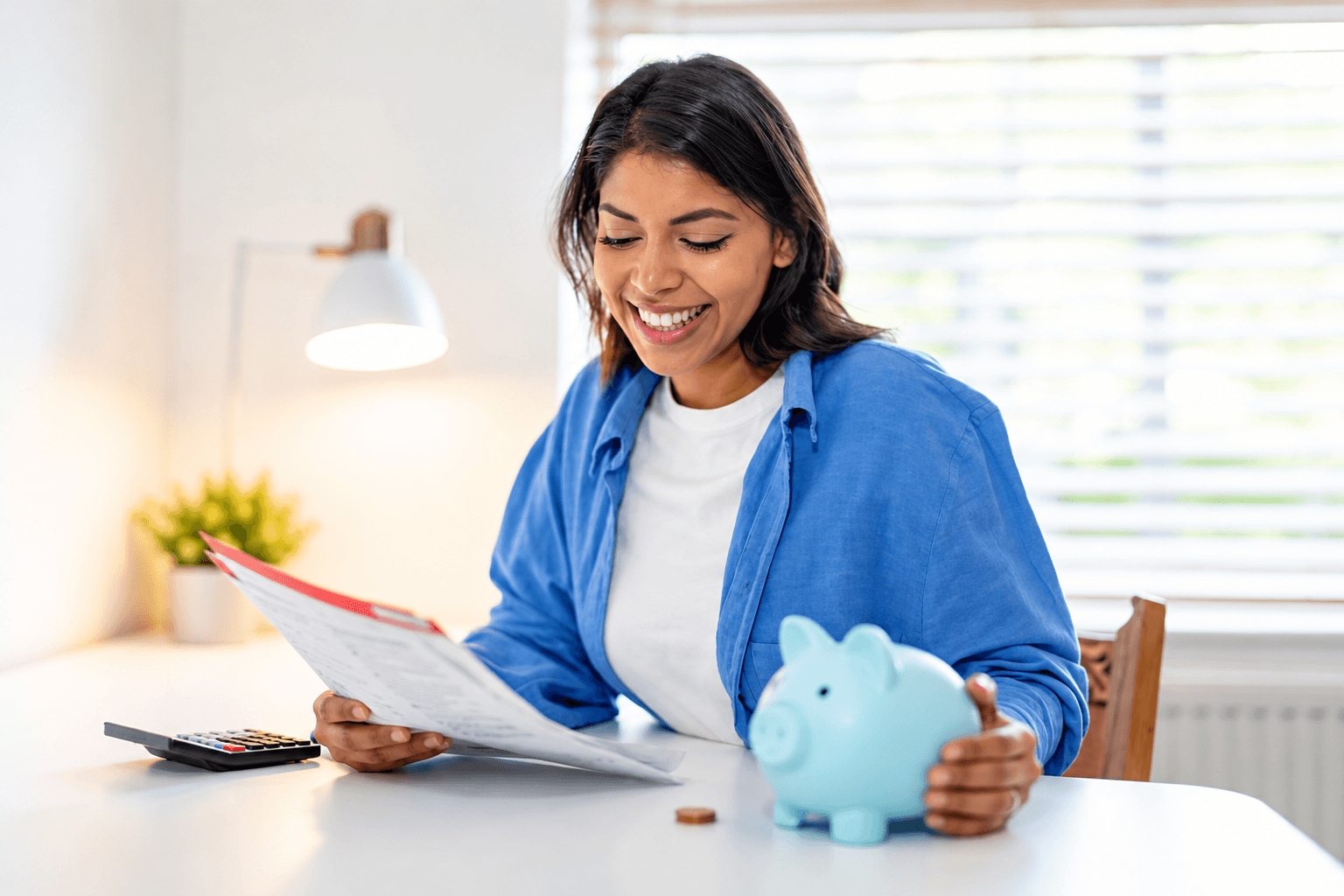 A smiling woman reviews financial paperwork at home with a calculator and piggy bank nearby. The image illustrates retirement savings planning and the transition from the Saver's Credit to the new Saver's Match, which offers eligible taxpayers government matching contributions beginning in 2027.