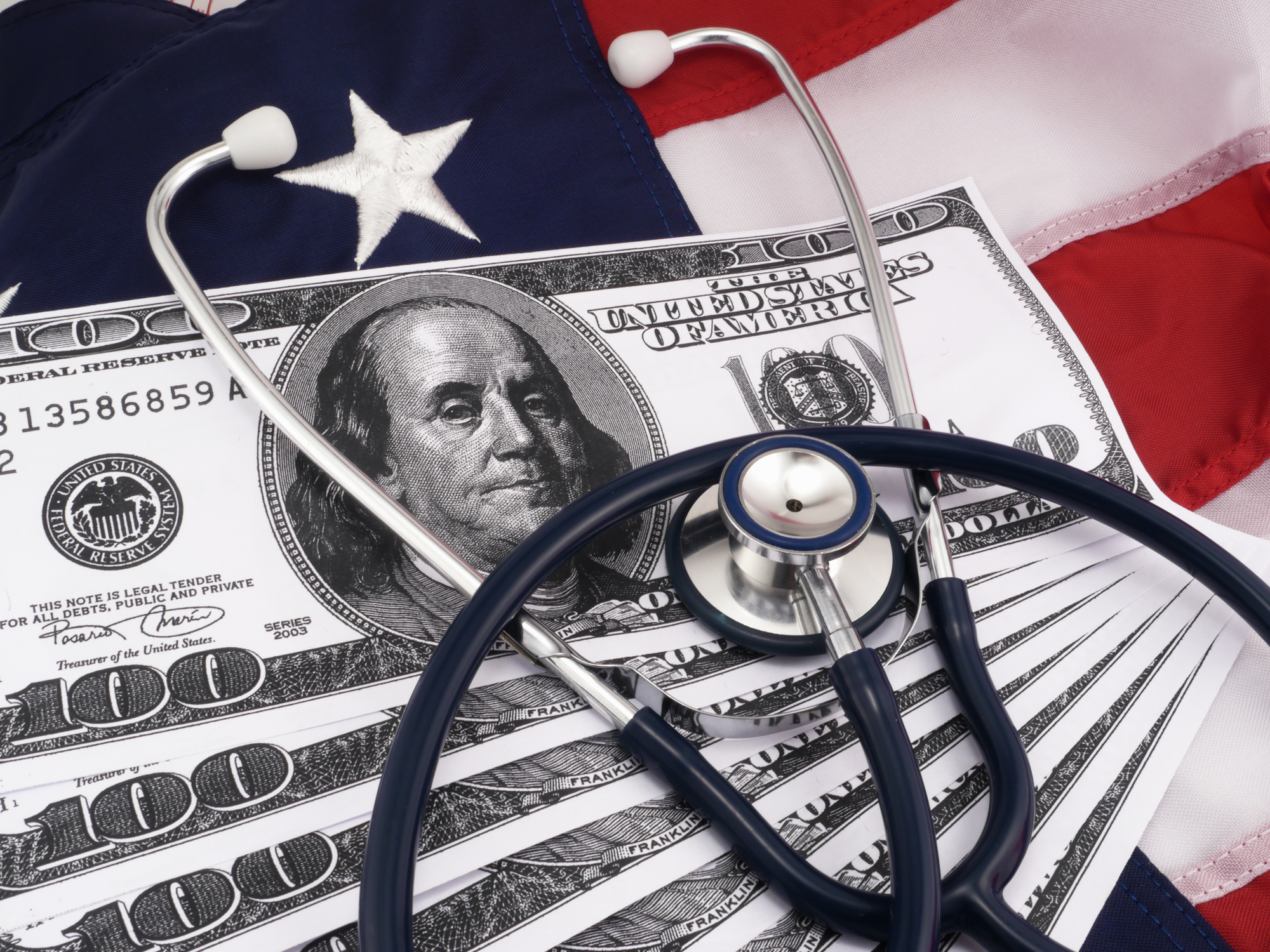 Stethoscope lying on a stack of one-hundred-dollar bills in front of an American flag, representing the growing crisis of medical debt in America.