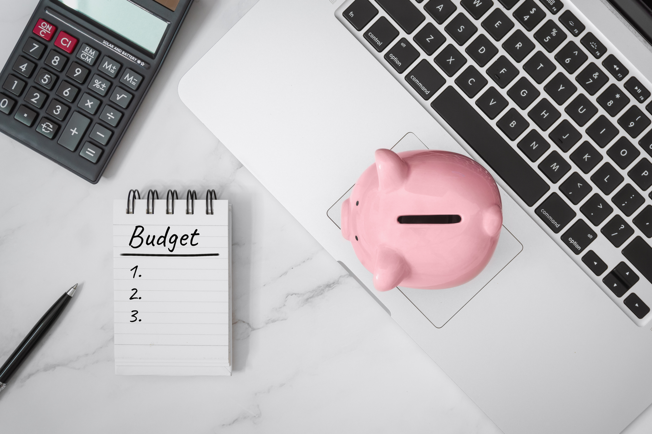 Budget calculator workspace with a piggy bank, calculator, laptop, and a handwritten budget checklist