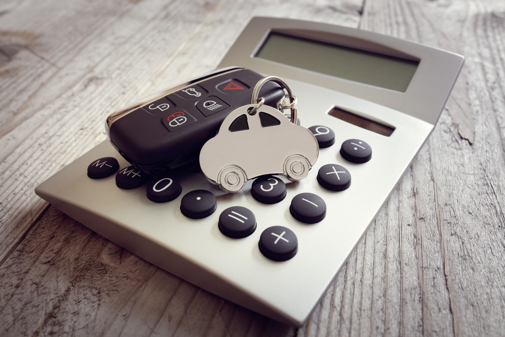 A car key fob and a silver car-shaped keychain resting on a silver digital calculator on a wooden table, representing a how much car can I afford calculator.